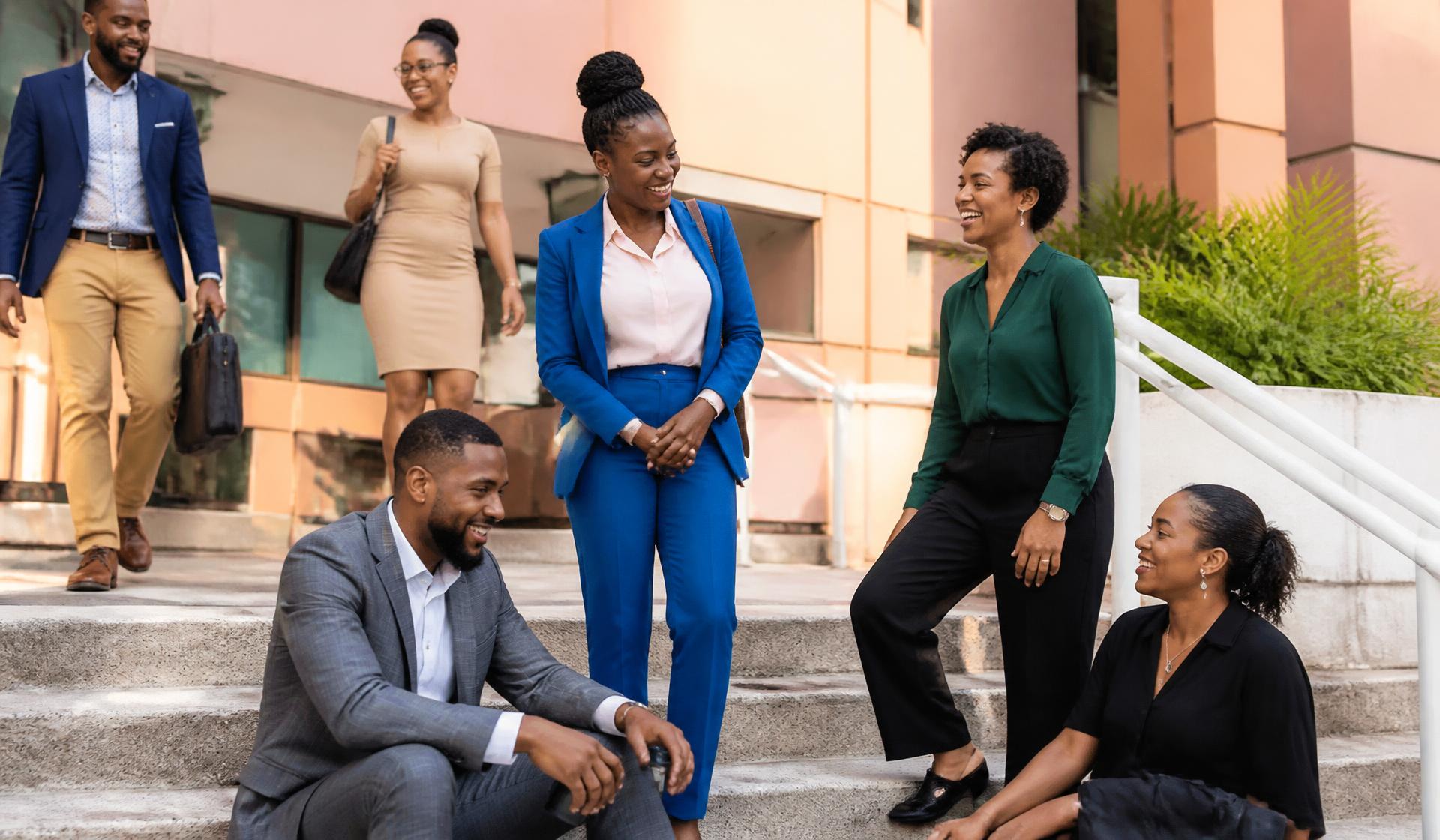 Jamaican professionals laughing together on the steps of a modern pastel Kingston building in late-afternoon light